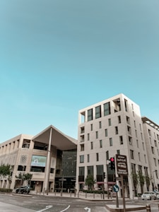 A modern architectural structure with clean lines and a mix of glass and stone elements is present. The building has multiple stories, featuring large windows and balconies. There is a prominent entrance to the Galleria, with trees and a clear blue sky in the background. Vehicles are present on the street, and there are traffic signs, including one in both English and Arabic indicating to look right for the tramway.