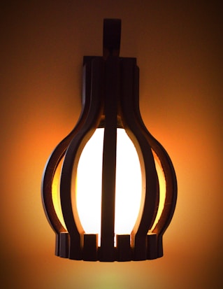 A warmly lit, modern wooden wall sconce is casting a soft glow against a plain background. The lamp has a unique, rounded design with evenly spaced wooden slats, creating a visually appealing silhouette.