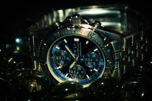 A silver chronograph wristwatch with a metallic band is surrounded by reflective glass spheres. The watch face is detailed with multiple subdials and bold hour markers, set against a dark backdrop that emphasizes its sleek design.