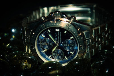 A silver chronograph wristwatch with a metallic band is surrounded by reflective glass spheres. The watch face is detailed with multiple subdials and bold hour markers, set against a dark backdrop that emphasizes its sleek design.