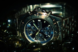 A silver chronograph wristwatch with a metallic band is surrounded by reflective glass spheres. The watch face is detailed with multiple subdials and bold hour markers, set against a dark backdrop that emphasizes its sleek design.