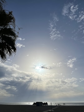 The image captures a serene beach scene under a bright sun with rays streaming through scattered clouds. Silhouettes of people are walking along the shoreline, and there is a cluster of beach huts or shaded areas in the distance. A palm tree is visible on the left edge of the image, adding to the tropical feel.
