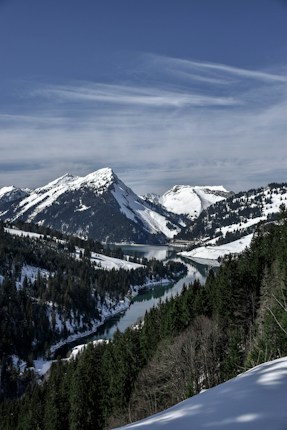 A serene mountainous landscape features snow-covered peaks under a clear blue sky. A winding river cuts through the valley, surrounded by dense pine forests.