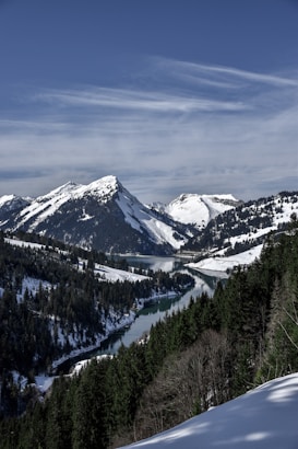 A serene mountainous landscape features snow-covered peaks under a clear blue sky. A winding river cuts through the valley, surrounded by dense pine forests.