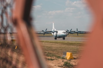A large turboprop aircraft is visible on the runway, framed through an orange-colored fence in the foreground. The sky above is partly cloudy, and the surrounding landscape appears to be grassy, typical of an airport environment. A yellow sign with a letter and number is also visible near the fence.