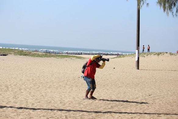 A photographer dressed in a red shirt and blue shorts is standing on a sandy beach. He is holding a camera with a long lens, aiming towards the sea. In the background, there are beach umbrellas lining the shore, with people enjoying the beach.