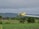 A yellow crop duster plane flying low over a green agricultural field, releasing a trail of spray. The landscape features lush greenery and a backdrop of dense trees and overcast, cloudy skies.