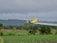 A yellow crop duster plane flying low over a green agricultural field, releasing a trail of spray. The landscape features lush greenery and a backdrop of dense trees and overcast, cloudy skies.