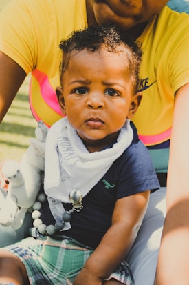 A baby with curly hair sits on someone's lap, wearing a navy blue shirt and plaid shorts. A white bib with text is around the baby's neck, and a stuffed toy is held by the baby. The person holding the baby is wearing a yellow shirt with a visible logo.
