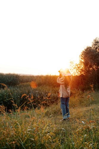A person stands in a grassy field, facing the sunset while holding a camera. The warm glow of the setting sun illuminates the scene, casting a golden hue over the landscape. Tall grasses and reeds surround the area, and the horizon is softly blurred, highlighting the tranquil atmosphere.