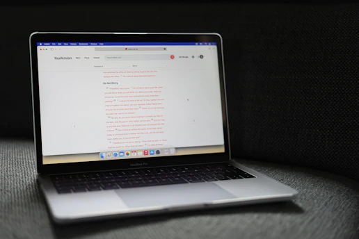 A MacBook Pro laptop displaying a webpage with a soft gray couch as a backdrop. The screen shows a website with text in the center, including a passage labeled 'Do Not Worry'. The laptop's backlit keyboard and touchpad are visible, and the bottom screen displays several application icons in the dock.