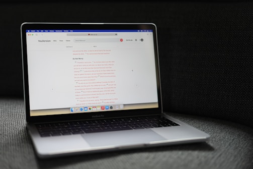A MacBook Pro laptop displaying a webpage with a soft gray couch as a backdrop. The screen shows a website with text in the center, including a passage labeled 'Do Not Worry'. The laptop's backlit keyboard and touchpad are visible, and the bottom screen displays several application icons in the dock.