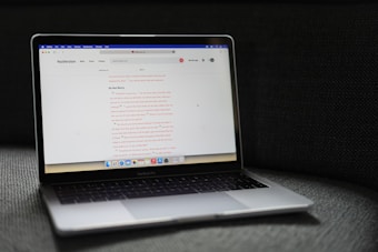 A MacBook Pro laptop displaying a webpage with a soft gray couch as a backdrop. The screen shows a website with text in the center, including a passage labeled 'Do Not Worry'. The laptop's backlit keyboard and touchpad are visible, and the bottom screen displays several application icons in the dock.