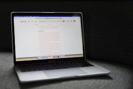A MacBook Pro laptop displaying a webpage with a soft gray couch as a backdrop. The screen shows a website with text in the center, including a passage labeled 'Do Not Worry'. The laptop's backlit keyboard and touchpad are visible, and the bottom screen displays several application icons in the dock.
