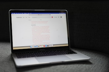 A MacBook Pro laptop displaying a webpage with a soft gray couch as a backdrop. The screen shows a website with text in the center, including a passage labeled 'Do Not Worry'. The laptop's backlit keyboard and touchpad are visible, and the bottom screen displays several application icons in the dock.