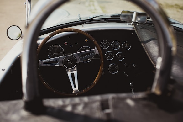 A vintage car interior featuring a wooden steering wheel and a dashboard with multiple round dials and gauges. The cockpit style design gives it a classic and elegant look.