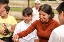 A group of people, including a woman in a red long-sleeve shirt, are gathered outdoors. They appear to be engaged in an activity that involves holding small objects. The woman is smiling and appears to be enjoying the moment. The background includes greenery and blurred figures.