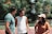 Three young adults are standing outdoors, engaged in conversation and laughter. Trees with green leaves and some orange hues form the background. The person on the left is wearing a teal t-shirt and sunglasses, the one in the middle is dressed in a white sleeveless shirt and a black cap, and the individual on the right is in a brown shirt and beige cap.