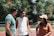Three young adults are standing outdoors, engaged in conversation and laughter. Trees with green leaves and some orange hues form the background. The person on the left is wearing a teal t-shirt and sunglasses, the one in the middle is dressed in a white sleeveless shirt and a black cap, and the individual on the right is in a brown shirt and beige cap.