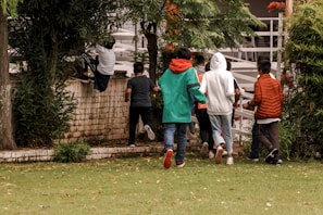 A group of seven young people, casually dressed in jeans and jackets, are seen in an outdoor setting surrounded by greenery. One person is climbing over a brick wall, while others appear to be following then through a gate. The scene suggests a playful or adventurous activity.