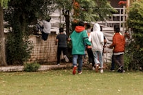 A group of seven young people, casually dressed in jeans and jackets, are seen in an outdoor setting surrounded by greenery. One person is climbing over a brick wall, while others appear to be following then through a gate. The scene suggests a playful or adventurous activity.