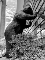 A large, geometric sculpture of a bear is seen peering into the windows of a modern building. The bear appears to be constructed with angular facets, creating a striking visual contrast. The scene is set in an urban environment with tall buildings and a cloudy sky.