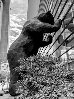A large, geometric sculpture of a bear is seen peering into the windows of a modern building. The bear appears to be constructed with angular facets, creating a striking visual contrast. The scene is set in an urban environment with tall buildings and a cloudy sky.