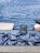 A pile of freshly caught fish is laid out on a flat surface by the water. Behind them, a small display advertises that various payment cards are accepted. The background shows a calm, blue body of water with several white birds floating in the distance.