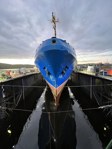 A large blue ship is docked in a narrow, industrial setting, with its bow prominently facing the viewer. The sky is overcast, and the setting appears to be a shipyard with various metallic structures and equipment surrounding the vessel.