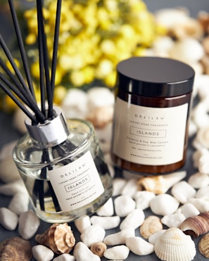 A luxury home fragrance set is displayed, consisting of a reed diffuser and a coconut and soy wax candle. The labels on both items read 'Delilah Islands.' They are surrounded by decorative white stones and seashells, with a blurred background of yellow flowers.