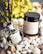 A luxury home fragrance set is displayed, consisting of a reed diffuser and a coconut and soy wax candle. The labels on both items read 'Delilah Islands.' They are surrounded by decorative white stones and seashells, with a blurred background of yellow flowers.