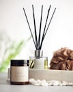 A reed diffuser and a coconut & soy wax candle labeled 'Serenity' are placed on a surface with white pebbles in front of them. A textured brown loofah is set to the side, with a blurred, soft green background featuring hints of foliage.