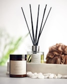 A reed diffuser and a coconut & soy wax candle labeled 'Serenity' are placed on a surface with white pebbles in front of them. A textured brown loofah is set to the side, with a blurred, soft green background featuring hints of foliage.