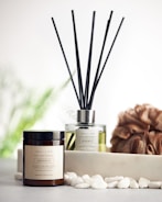 A reed diffuser and a coconut & soy wax candle labeled 'Serenity' are placed on a surface with white pebbles in front of them. A textured brown loofah is set to the side, with a blurred, soft green background featuring hints of foliage.