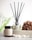 A reed diffuser and a coconut & soy wax candle labeled 'Serenity' are placed on a surface with white pebbles in front of them. A textured brown loofah is set to the side, with a blurred, soft green background featuring hints of foliage.