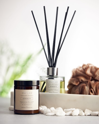 A reed diffuser and a coconut & soy wax candle labeled 'Serenity' are placed on a surface with white pebbles in front of them. A textured brown loofah is set to the side, with a blurred, soft green background featuring hints of foliage.
