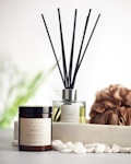 A reed diffuser and a coconut & soy wax candle labeled 'Serenity' are placed on a surface with white pebbles in front of them. A textured brown loofah is set to the side, with a blurred, soft green background featuring hints of foliage.