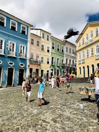 A vibrant street scene featuring colonial-style buildings painted in pastel colors such as blue, yellow, green, and pink. A group of people, including a man taking a selfie, are walking on a cobblestone street, while a large black bird flies overhead. Flags hang from one of the buildings, and outdoor stalls with colorful items are visible.