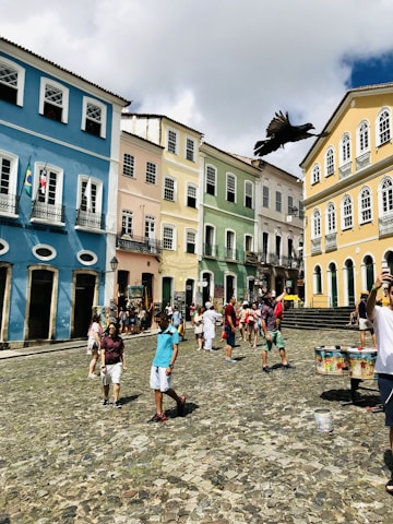 A vibrant street scene featuring colonial-style buildings painted in pastel colors such as blue, yellow, green, and pink. A group of people, including a man taking a selfie, are walking on a cobblestone street, while a large black bird flies overhead. Flags hang from one of the buildings, and outdoor stalls with colorful items are visible.