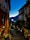 A picturesque narrow street at twilight, flanked by rustic buildings with shuttered windows. Dimly lit streetlights cast a warm glow on the cobblestone path. People sit and dine outside a quaint wine bar, while others walk down the street. In the background, a serene view of a lake and darkened hills is visible under the deep blue sky.
