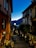 A picturesque narrow street at twilight, flanked by rustic buildings with shuttered windows. Dimly lit streetlights cast a warm glow on the cobblestone path. People sit and dine outside a quaint wine bar, while others walk down the street. In the background, a serene view of a lake and darkened hills is visible under the deep blue sky.