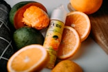A bottle of flavored liquid is surrounded by fresh fruits, including sliced and whole oranges and a halved mango. The liquid bottle features a label with a design showcasing fruit illustrations, and the composition is set on a light-colored surface with a wooden board in the background.