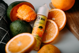 A bottle of flavored liquid is surrounded by fresh fruits, including sliced and whole oranges and a halved mango. The liquid bottle features a label with a design showcasing fruit illustrations, and the composition is set on a light-colored surface with a wooden board in the background.