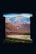 Through the viewfinder of a camera, a landscape featuring rugged mountains in shades of blue and orange appears. A riverbed or stream lined with patches of green vegetation runs through the arid terrain, adding contrast to the earthy tones of the hills.