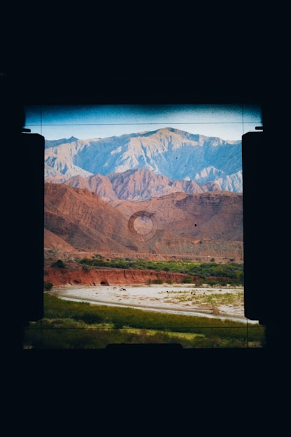 Through the viewfinder of a camera, a landscape featuring rugged mountains in shades of blue and orange appears. A riverbed or stream lined with patches of green vegetation runs through the arid terrain, adding contrast to the earthy tones of the hills.