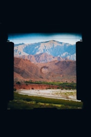 Through the viewfinder of a camera, a landscape featuring rugged mountains in shades of blue and orange appears. A riverbed or stream lined with patches of green vegetation runs through the arid terrain, adding contrast to the earthy tones of the hills.