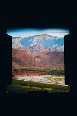 Through the viewfinder of a camera, a landscape featuring rugged mountains in shades of blue and orange appears. A riverbed or stream lined with patches of green vegetation runs through the arid terrain, adding contrast to the earthy tones of the hills.