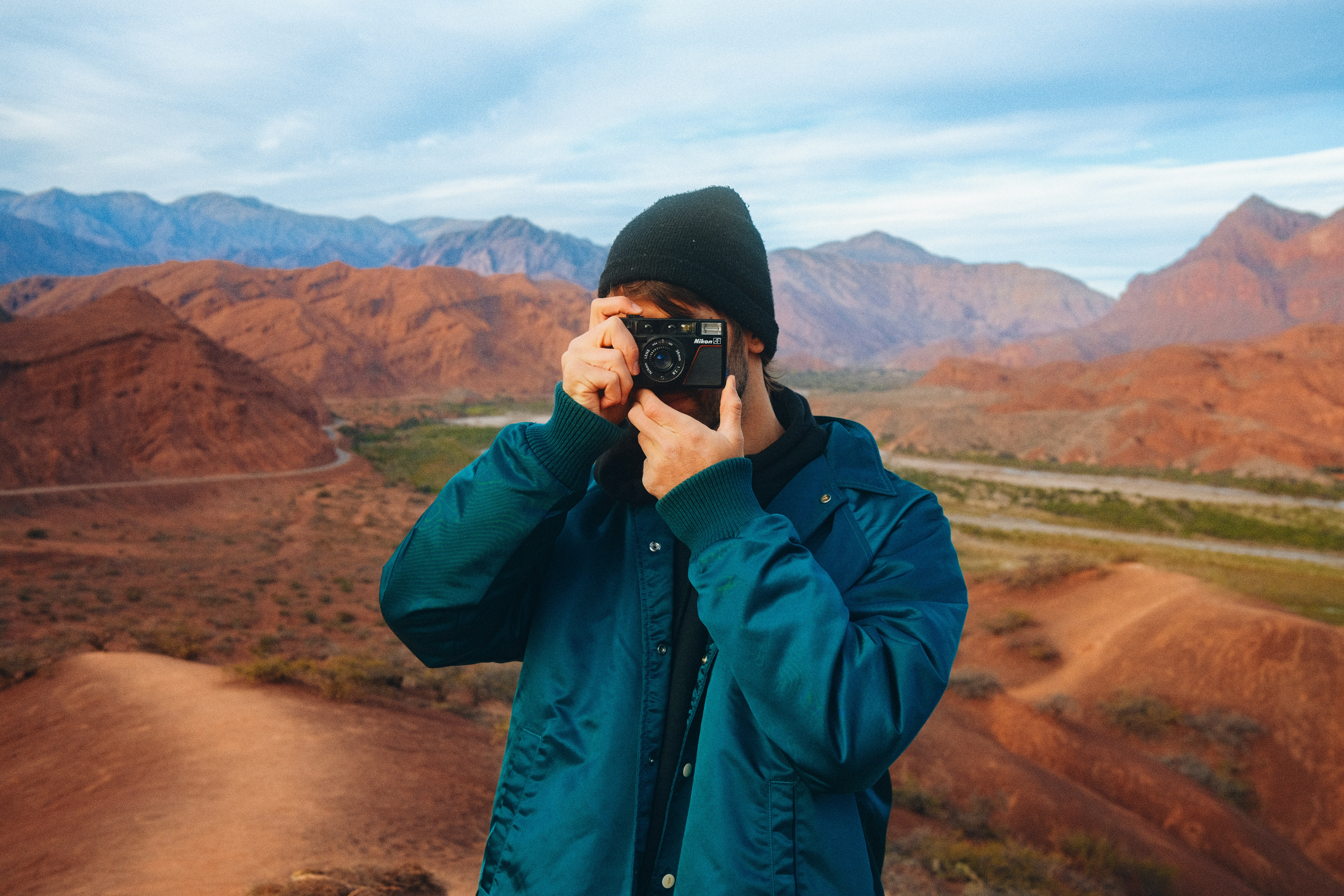 a man holding a camera, Portrait of a film photographer with a Nikon camera, blue jacket and warm clothes. Behind is an immense valley landscape with mountains, rivers and vegetation with a road to the side.
