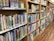 A library shelf filled with colorful children's books organized neatly. A variety of books with vivid covers and titles are visible, alongside some upright display books and a holder with pencils.