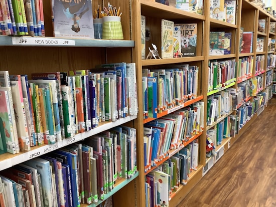 A library shelf filled with colorful children's books organized neatly. A variety of books with vivid covers and titles are visible, alongside some upright display books and a holder with pencils.